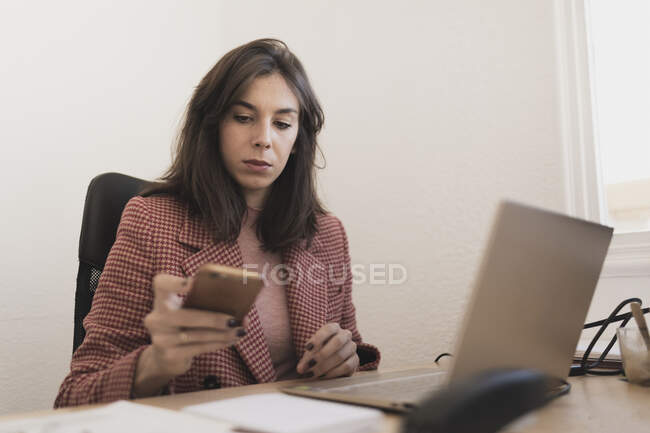 Jeune femme concentrée utilisant un téléphone portable et assise à table avec des moniteurs au bureau — Photo de stock