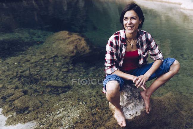 Happy adult woman sitting on rock in tranquil transparent water of lake enjoying nature and smiling away — Stock Photo