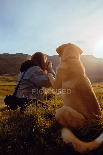 Вид ззаду молода жінка знімає пейзаж на камеру біля смішного і доброзичливого собаки між лугом і блакитним небом — стокове фото