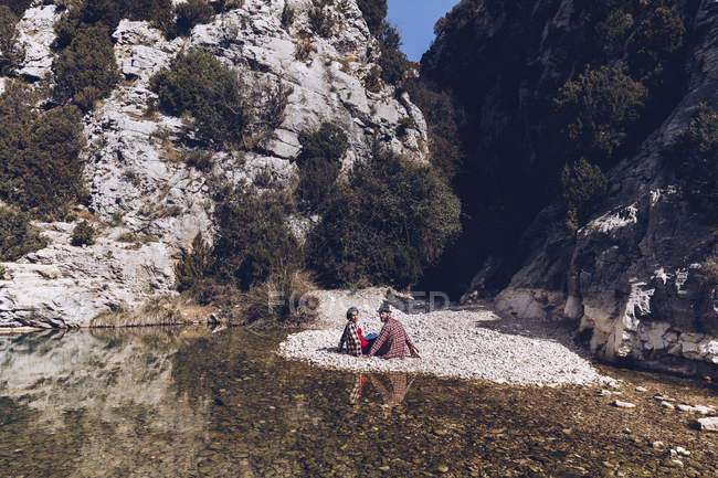 Vista lateral de pareja joven sentada en la costa rocosa del río de montaña cerca del acantilado - foto de stock