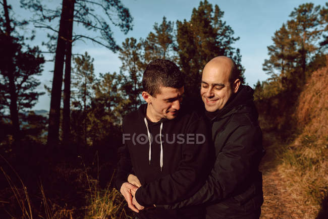 Cheerful homosexual couple embracing on walkway in forest in sunny day on blurred background — Stock Photo