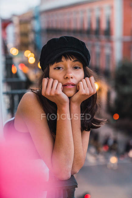 Young woman in cap looking at camera and standing on balcony near street with lights in evening — Stock Photo
