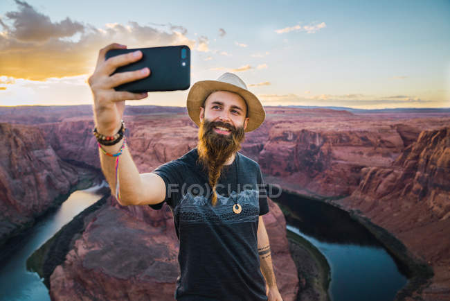Hombre con sombrero sonriendo y tomando selfie mientras está parado contra magnífico cañón y río durante el atardecer en la costa oeste de EE.UU. - foto de stock