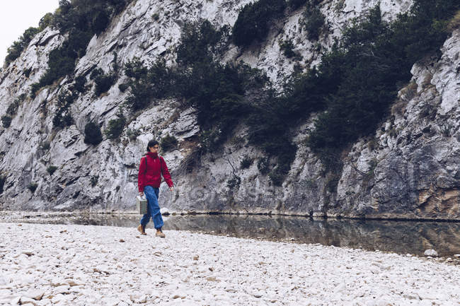 Mulher casual transportando caso e andando na costa de fluxo claro de água na natureza — Fotografia de Stock