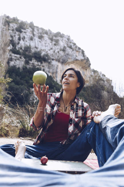 Hombre acostado en cuadros con la mujer sentada cerca y arrojando manzana disfrutando de tiempo juntos en el picnic en la naturaleza - foto de stock