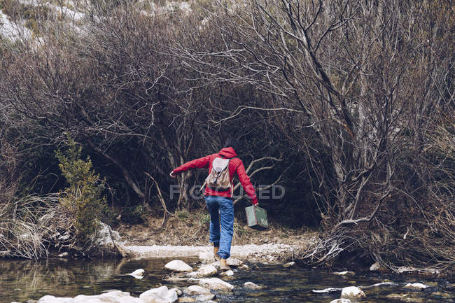 Back view of casual woman carrying case and jumping on rocks of clear stream of water in nature — Stock Photo