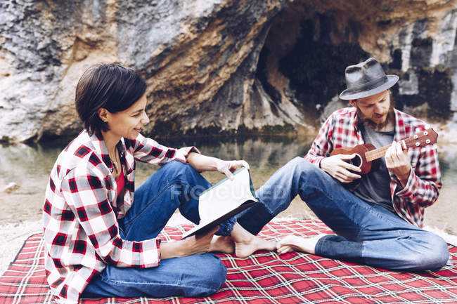 Adult man and woman on checkered plaid with book and small ukulele having picnic on lake shore — Stock Photo