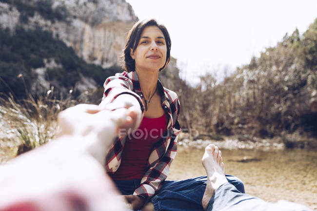Hombre de la cosecha cogido de la mano de la mujer adulta sonriente mientras está sentado en la costa del lago a la luz del sol - foto de stock