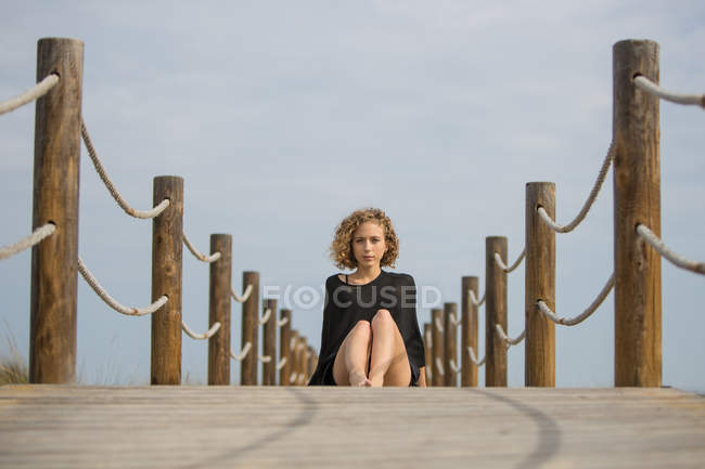 Молодая серьезная женщина сидит на деревянном пешеходном мосту и смотрит в камеру — стоковое фото