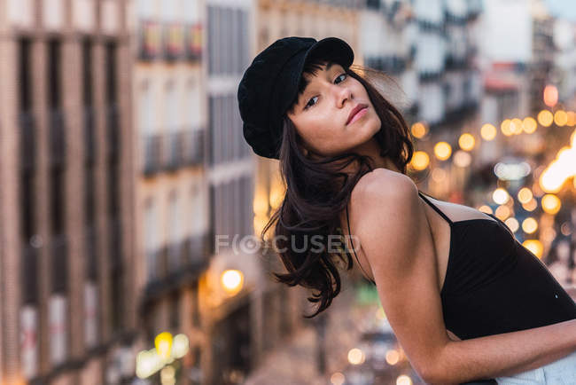 Young slim woman in cap looking at camera and standing on balcony on street with lights in evening — Stock Photo