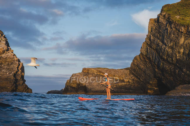 Frau in Badebekleidung steht auf dem Paddelbrett und rudert mit dem Paddel auf der Oberfläche des gewellten Meerwassers gegen den bewölkten Himmel — Stockfoto