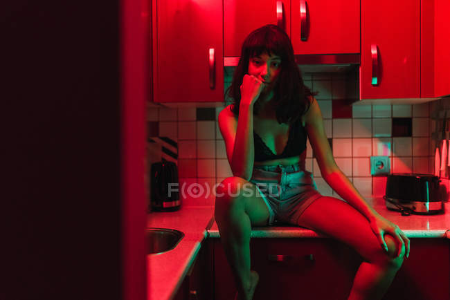 Sensual joven morena mujer en lencería mirando a la cámara y sentado en la cocina entre el enrojecimiento - foto de stock