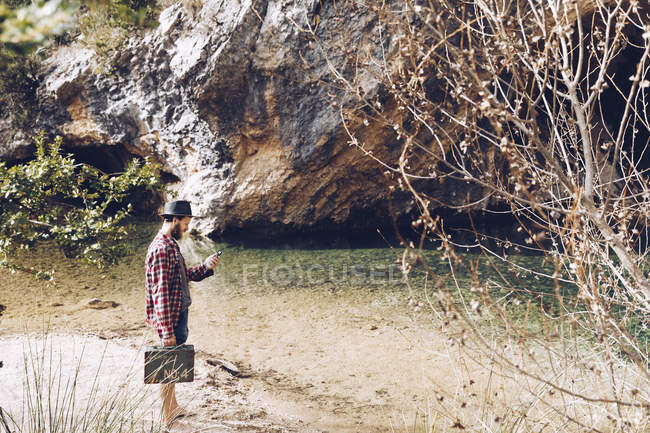 Vista lateral do homem viajante segurando pequena caixa e usando telefone enquanto está em pé na costa arenosa de lago tranquilo contra penhasco — Fotografia de Stock