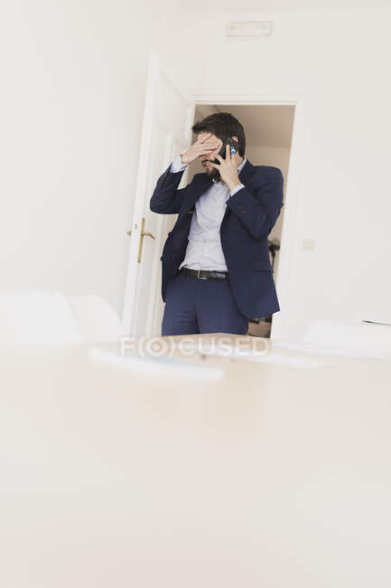 Hombre joven concentrado con la mano en el bolsillo hablando por teléfono móvil en la habitación con sillas y mesa - foto de stock