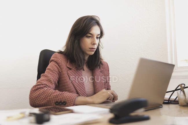 Jeune femme concentrée lisant des documents et assise à table avec ordinateur portable et smartphone au bureau — Photo de stock