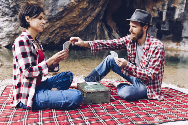 Side view of man and woman in plaid shirts playing cards on plaid having picnic on shore of lake in cliffs — Stock Photo