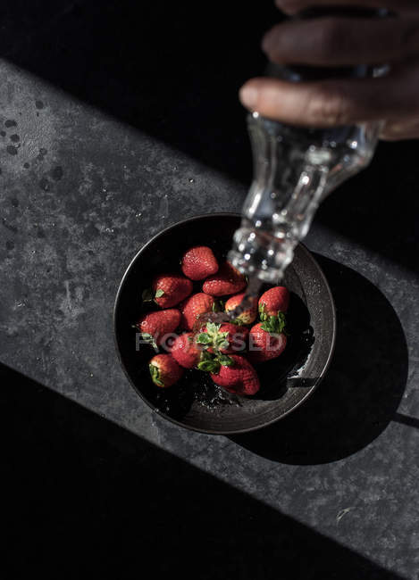 Mão cortada de pessoa segurando garrafa e derramando água de morangos frescos em tigela no fundo escuro — Fotografia de Stock