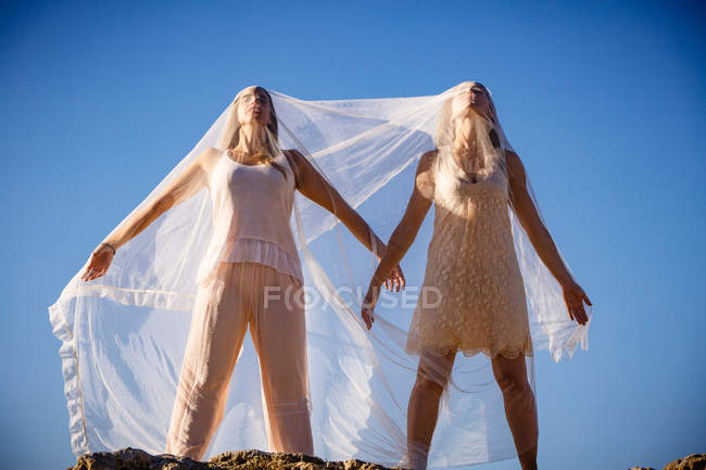Von unten junge geheimnisvolle Frauen mit erhobenen Händen, die weißes Textil halten und auf Felsen und blauem Himmel posieren — Stockfoto