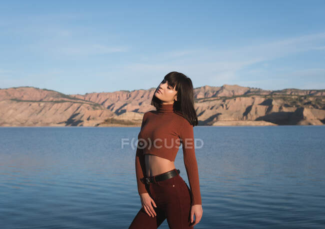 Attraktive schlanke Frau in stylischem Outfit, die die Augen geschlossen hält, während sie gegen ruhiges Wasser und atemberaubende Hügel in der Natur steht — Stockfoto