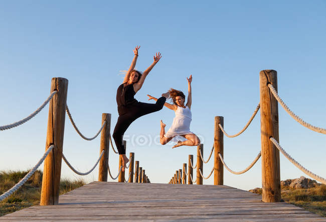Young ballerinas in white and black wear with upped leg in air on footbridge and blue sky in sunny day — Stock Photo