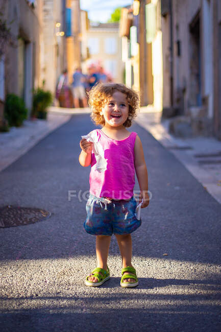 Menina engraçada feliz limpar a boca com guardanapo enquanto está em pé na rua de asfalto entre edifícios no verão — Fotografia de Stock
