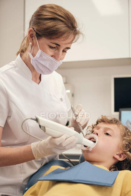 Woman in mask and doctor uniform making scan of teeth of little boy while working in dentist clinic — Stock Photo