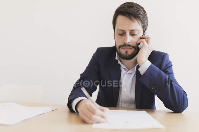 Giovane maschio concentrato che parla sul telefono cellulare e tiene la penna vicino ai documenti a tavola in ufficio — Foto stock