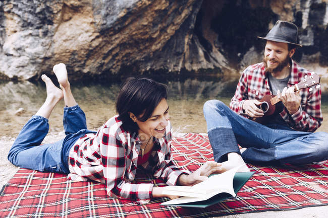 Hombre y mujer adultos en cuadros a cuadros con libro y pequeño ukelele teniendo picnic en la orilla del lago - foto de stock