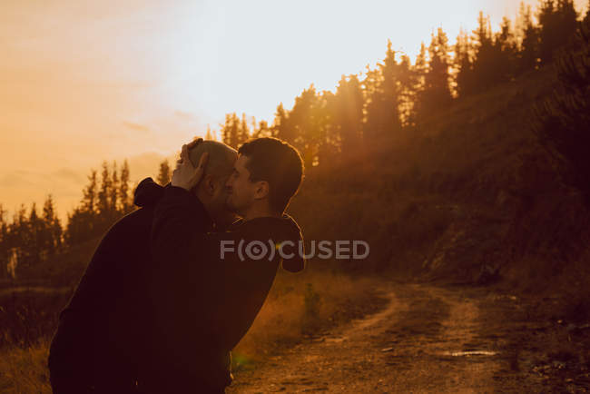 Feliz pareja homosexual abrazándose y besándose en el camino en el bosque en un día soleado - foto de stock