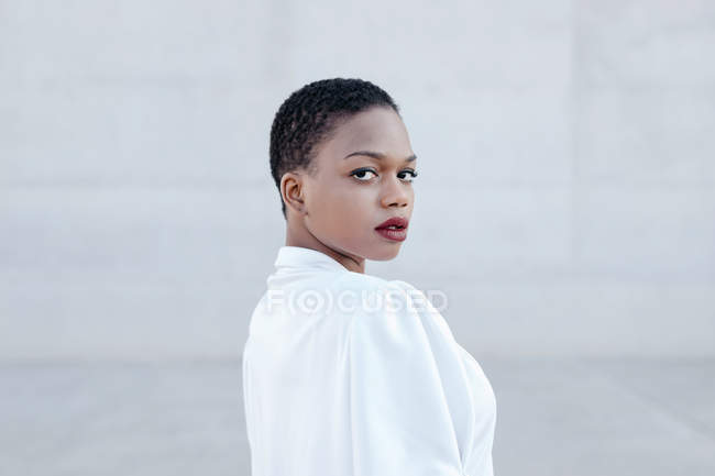 Moda mulher étnica de cabelos curtos em camisa branca posando contra a parede cinza — Fotografia de Stock