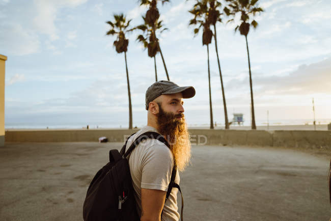 Vista lateral del tipo barbudo con mochila mirando a la cámara mientras está de pie cerca de la playa con palmera - foto de stock