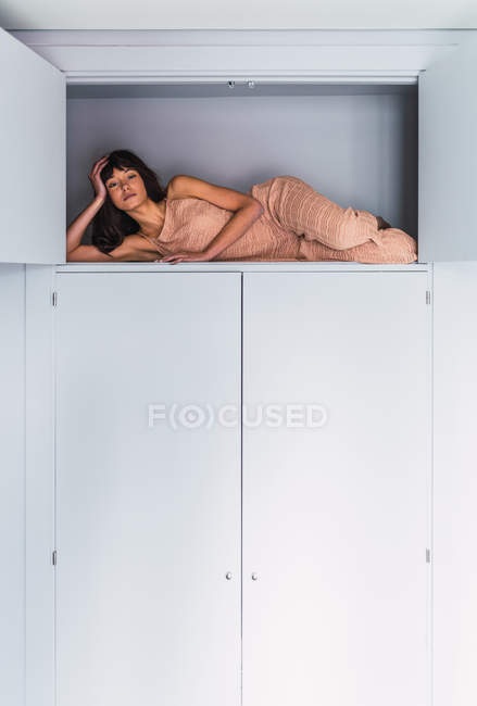 Ritratto di giovane donna sdraiata sul ripiano alto dell'armadio in camera da letto — Foto stock