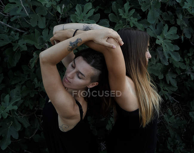Side view of attractive young women with closed eyes and upped hands standing near green shrub — Stock Photo