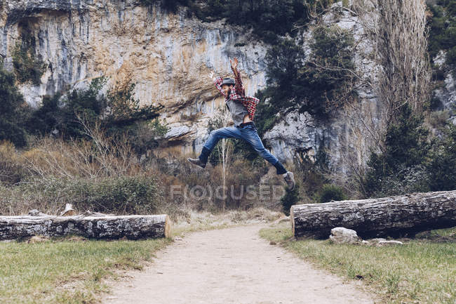 Homme décontracté sautant parmi les troncs tombés sur la route rurale contre une belle falaise — Photo de stock