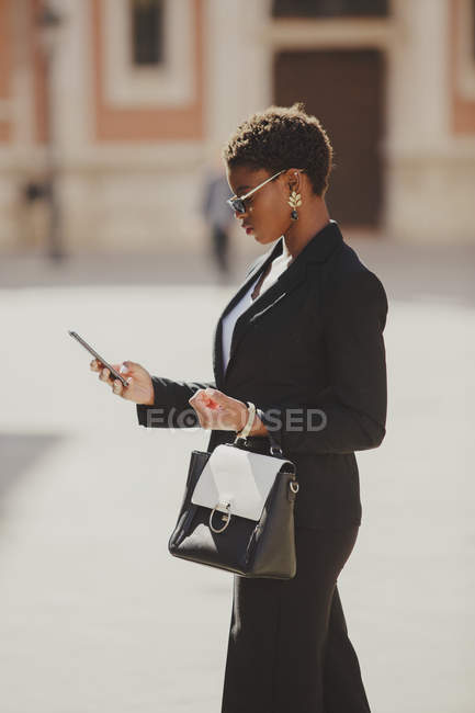 Mujer elegante afroamericana segura en traje y gafas de sol con bolsa y teléfono móvil en la calle - foto de stock