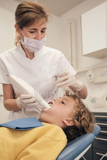 Женщина в маске и униформе врача делает сканирование зубов маленького мальчика во время работы в стоматологической клинике — стоковое фото