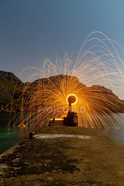 Long exposure of silhouette of human with burning round and fireworks on pier near water and mountains in evening — Stock Photo