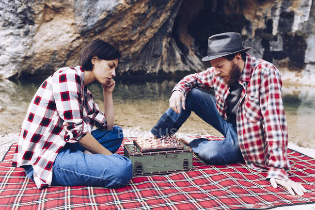 Side view of man and woman in plaid shirts playing chess on plaid having picnic on shore of lake in cliffs — Stock Photo