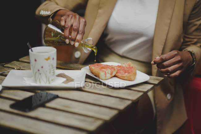 Mãos de mulher elegante derramando óleo em sanduíches e sentado à mesa com xícara de bebida e telefone celular no café da rua — Fotografia de Stock