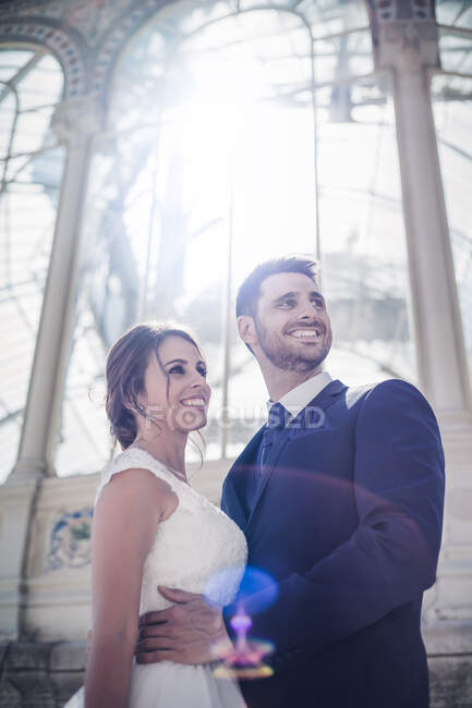 Знизу молодий елегантний чоловік приймає жінку у весільній сукні біля ретро-палацу з багатьма вікнами в сонячний день — стокове фото