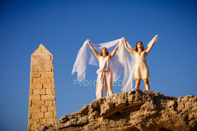 Dal basso giovani donne misteriose con le mani alzate in possesso di tessuti bianchi e in posa su rocce e cielo blu — Foto stock