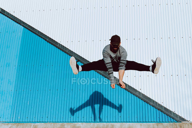 Bearded guy in trendy outfit leaping up and looking at camera against blue wall of modern building on sunny day — Stock Photo