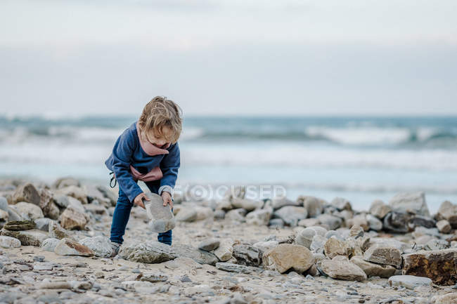 Niño positivo sosteniendo roca cerca de montón de piedras en la costa cerca de mar ondulante sobre fondo borroso - foto de stock