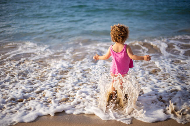 Back view of funny girl on sand shore going in water with foam and splashes in summer — Stock Photo