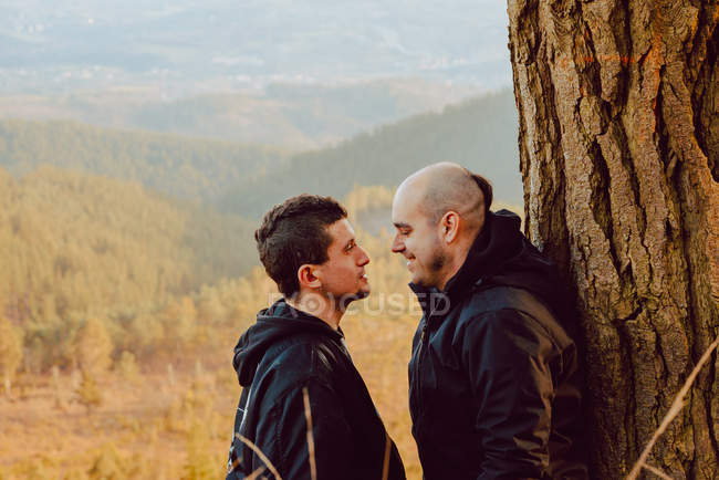 Vista lateral do casal homossexual alegre olhando um para o outro perto da árvore na floresta e vista pitoresca do vale — Fotografia de Stock