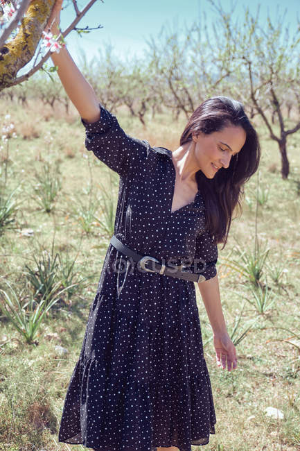 Jeune femme réfléchie en robe debout dans le jardin avec des arbres fruitiers en fleurs et branche exploitation — Photo de stock
