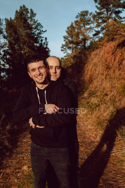 Portrait of cheerful homosexual couple embracing in forest in sunny day on blurred background — Stock Photo