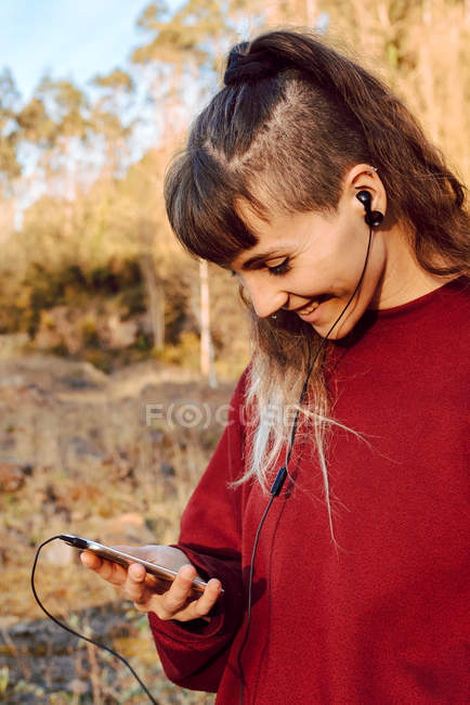 Молодая хипстерша с пирсингом и наушниками слушает музыку с мобильного телефона в сельской местности — стоковое фото