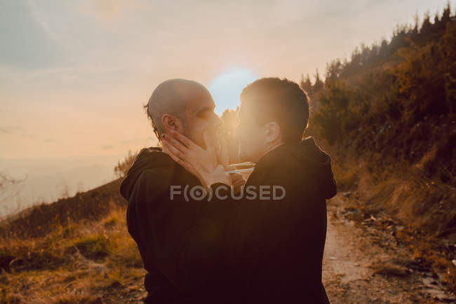 Romântico casal homossexual abraçando no caminho em montanhas em luz solar brilhante ao pôr do sol — Fotografia de Stock