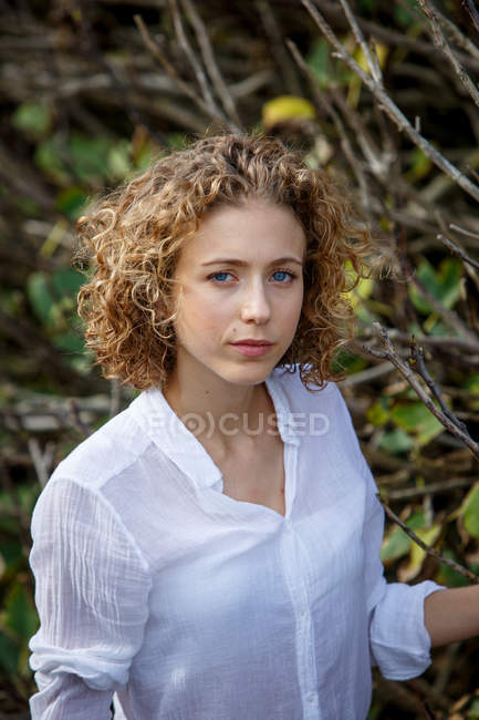 Jovem mulher pensativa posando perto de ramos secos de arbusto no fundo embaçado — Fotografia de Stock